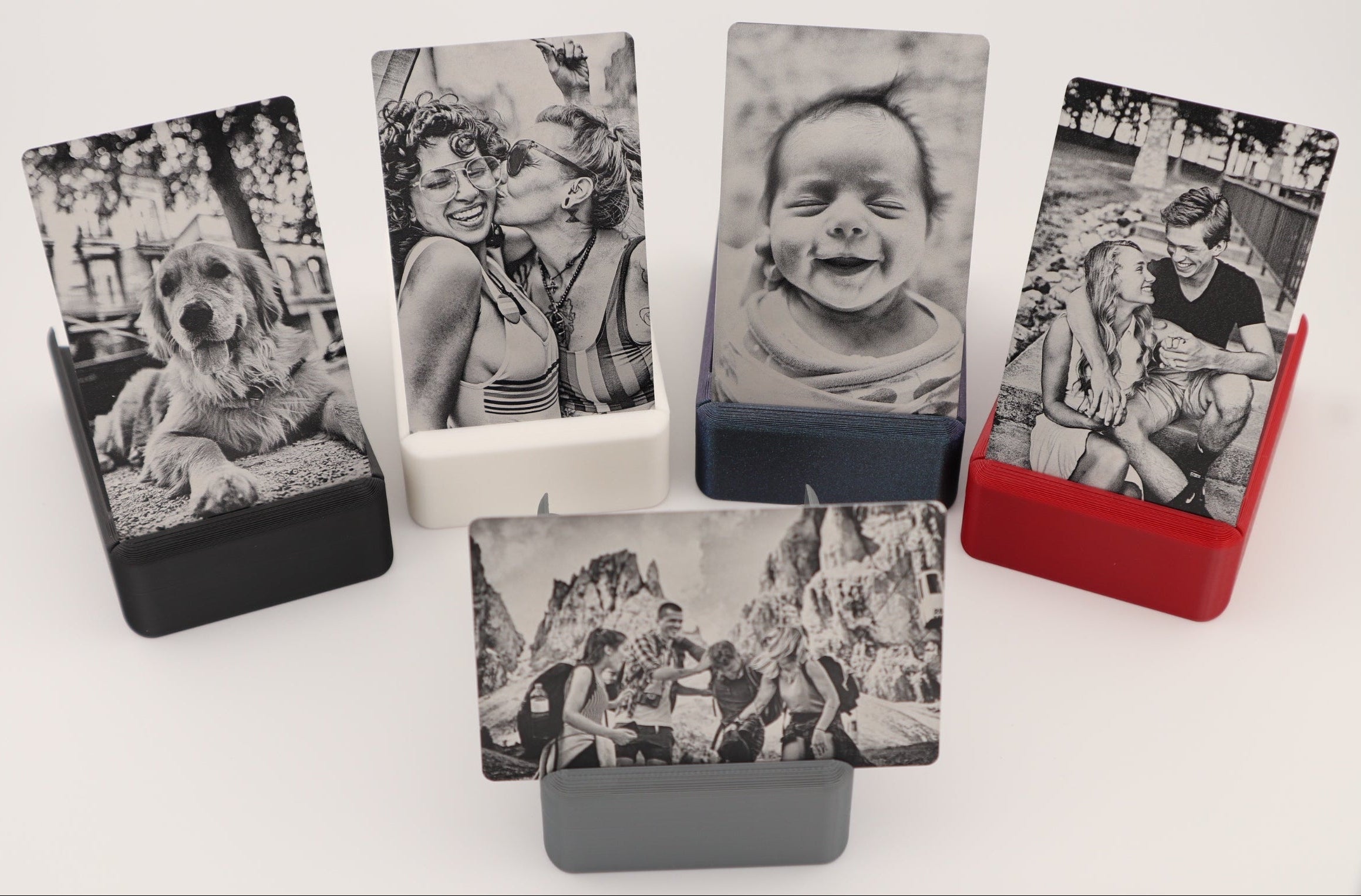 Five custom photo cards in various colors (black, white, navy, red, gray) displayed in matching rectangular holders on a clean white background. Each card features a different black and white photograph, including a dog, a couple, a baby, and outdoor scenes.