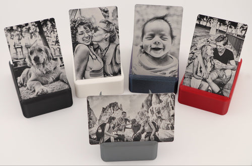 Five custom photo cards in various colors (black, white, navy, red, gray) displayed in matching rectangular holders on a clean white background. Each card features a different black and white photograph, including a dog, a couple, a baby, and outdoor scenes.
